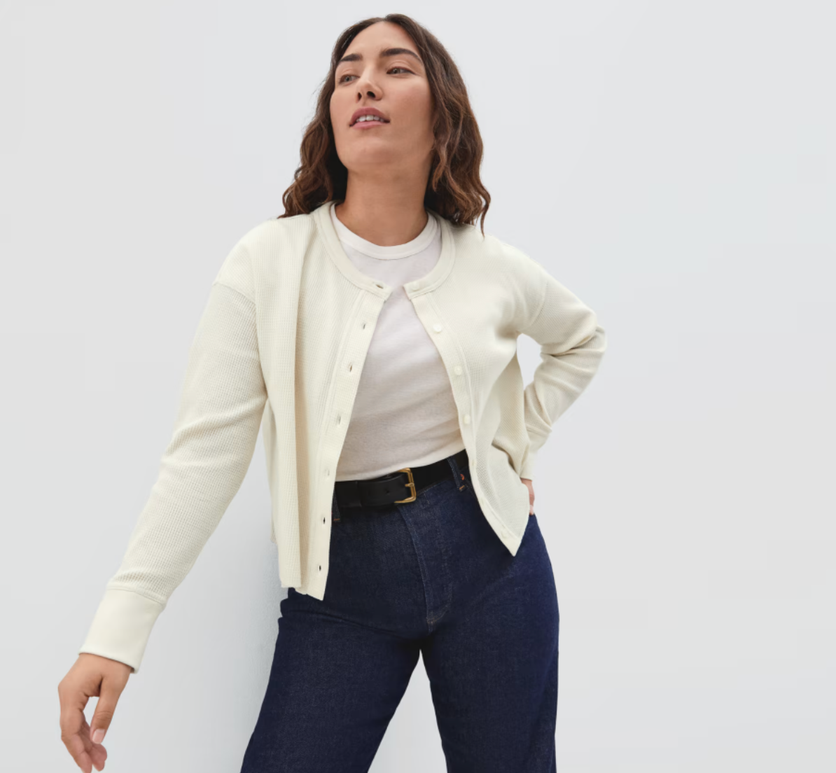 Model is wearing a light cream cardigan over a white top and denim jeans