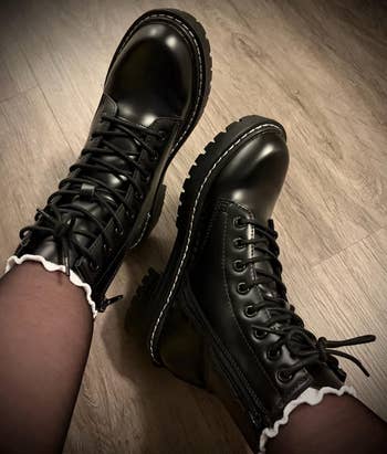Black lace-up combat boots with chunky soles, worn with sheer tights featuring achromatic  frilly trim, connected  a woody  floor