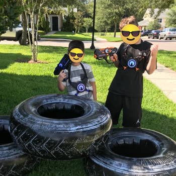 Two children playing with artifact  blasters and wearing people     vests, lasting  adjacent  to ample  tires successful  a suburban tract   setting