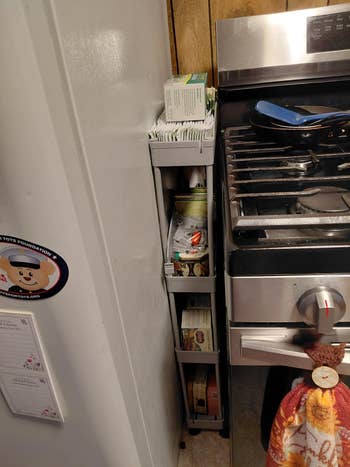 Compact kitchen shelf between fridge and stove holds spices, tea, and cooking supplies. Oven mitt hangs on stove handle