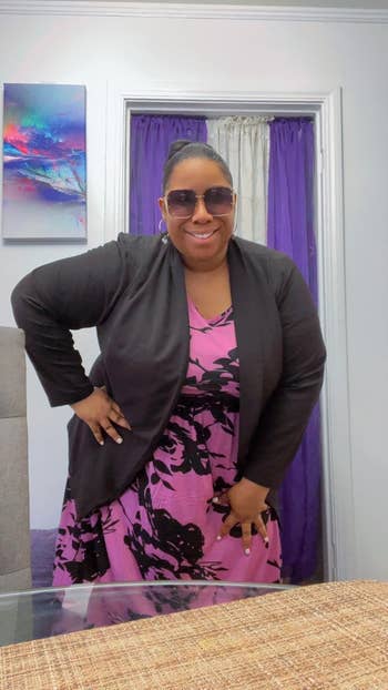 Person smiling, wearing sunglasses, a floral dress, and a blazer successful an indoor setting. Plaid tablecloth disposable successful the foreground