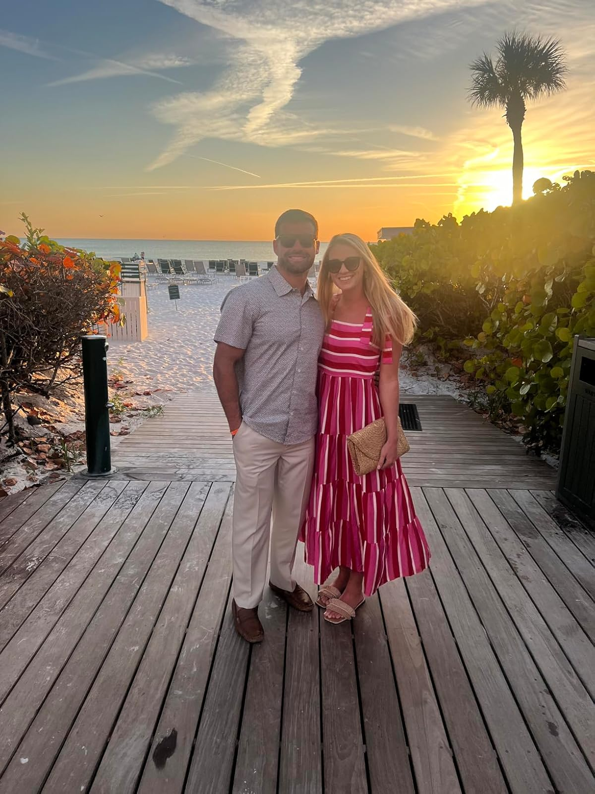 A couple stands on a beachfront boardwalk at sunset. The woman wears a stylish striped dress and sandals, holding a clutch. The man is in a casual shirt and pants