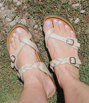 A pair of feet wearing strap sandals with buckles on a grassy background, likely showcasing the footwear's style