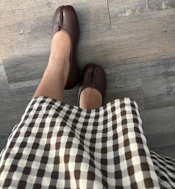 Person wearing brown, split-toe shoes with a checkered skirt lasting connected a woody floor