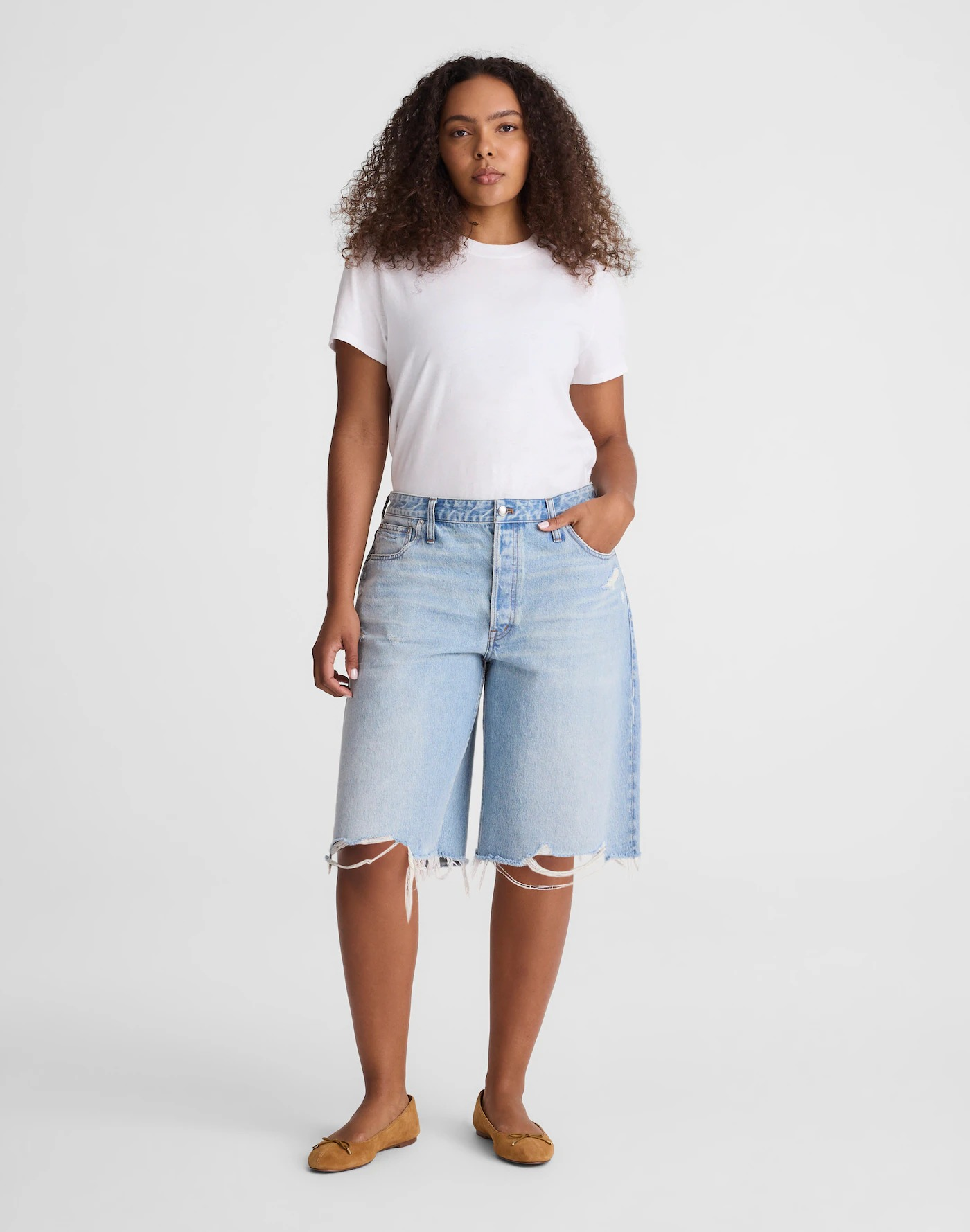 Person wearing a plain t-shirt, frayed denim shorts, and flat shoes, standing against a neutral background
