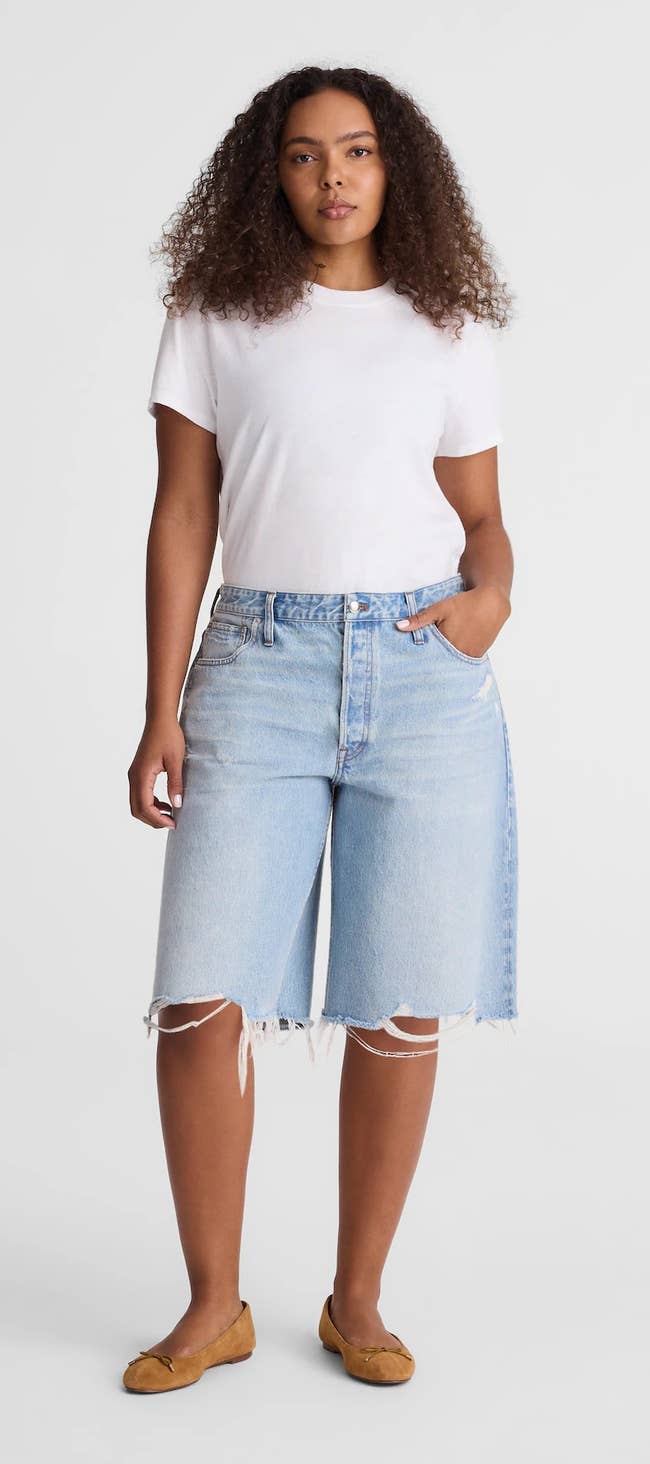 Person wearing a plain t-shirt, frayed denim shorts, and flat shoes, standing against a neutral background