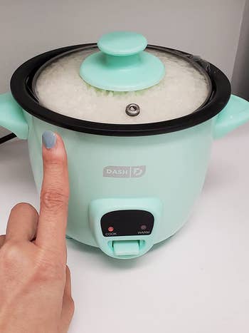 reviewer holding their finger up next to the teal rice cooker for scale