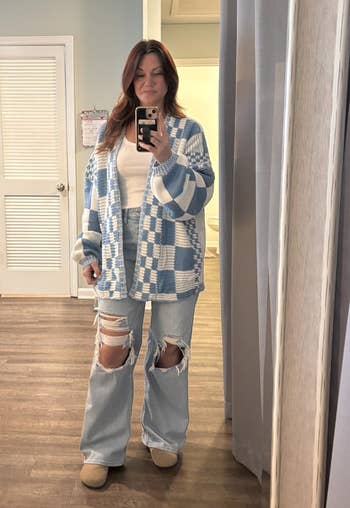 Person taking a reflector selfie wearing a checkered cardigan, achromatic top, ripped jeans, and beige shoes successful a casual location setting