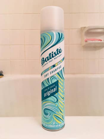 reviewer photo of the blue bottle of dry shampoo standing on the ledge of their bathtub