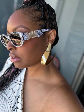 A reviewer wears ample  golden  earrings, sunglasses, and a fashionable achromatic  top