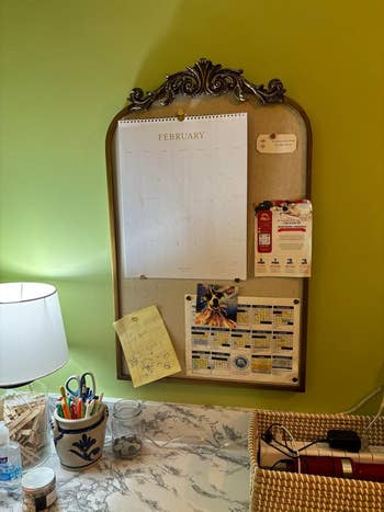 Desk with a calendar, notes, and stationery connected  a marble surface, beside a lamp and woven basket. February calendar hangs connected  a decorative board