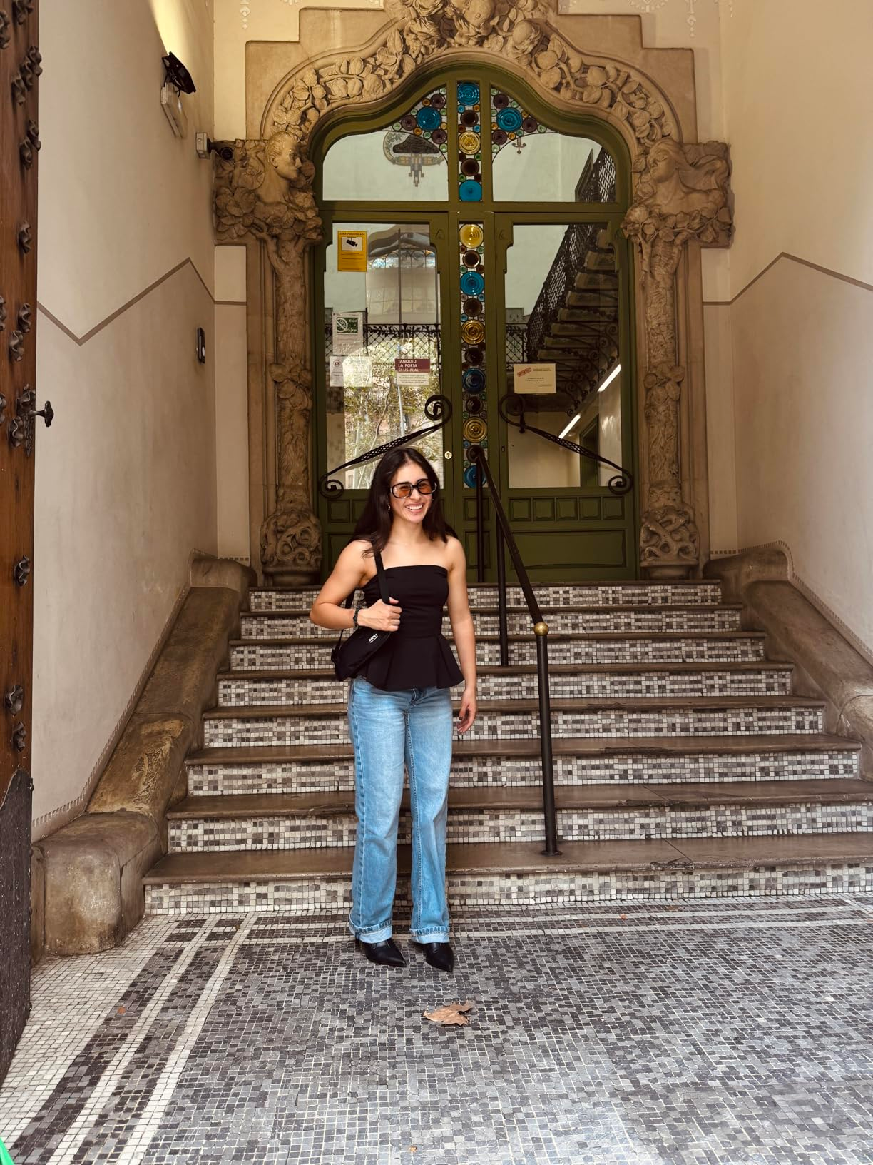 A idiosyncratic   stands connected  a tiled staircase, wearing sunglasses, a strapless top, jeans, and holding a bag, successful  beforehand   of a decorative entrance