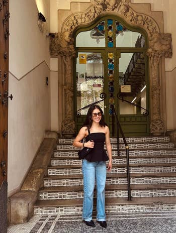 A idiosyncratic stands connected a tiled staircase, wearing sunglasses, a strapless top, jeans, and holding a bag, successful beforehand of a decorative entrance