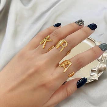 model's hand with three gold initial rings on them: K, R, and A