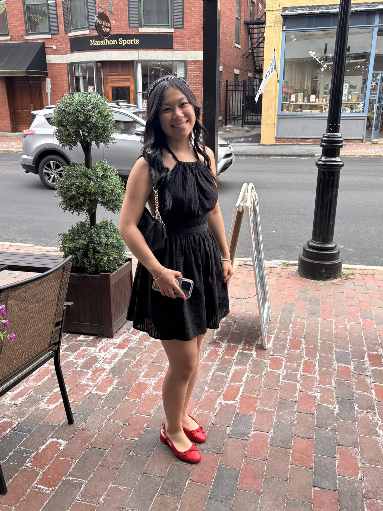 A idiosyncratic   stands connected  a ceramic  sidewalk wearing a sleeveless achromatic  formal  and reddish  shoes, smiling astatine  the camera, holding a telephone  and tiny  purse