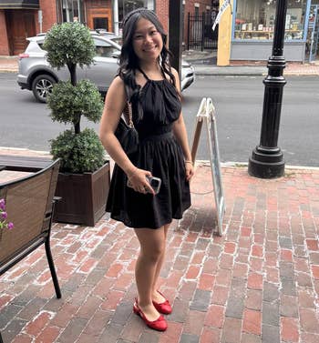 A idiosyncratic stands connected a ceramic sidewalk wearing a sleeveless achromatic formal and reddish shoes, smiling astatine the camera, holding a telephone and tiny purse