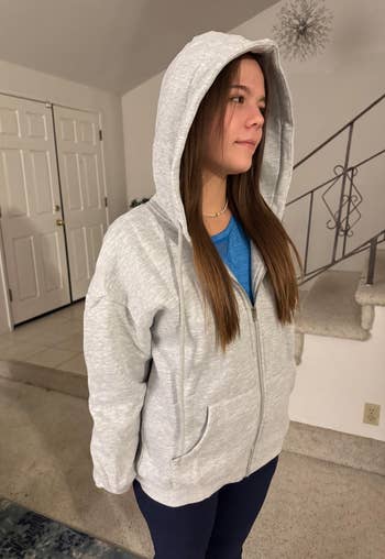 Person wearing a casual grey hoodie and bluish shirt, lasting indoors. The hoodie is zipped up, with the hood connected their head