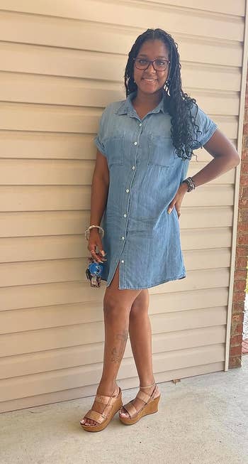 A person stands outside, smiling, in a buttoned denim dress with ruffle sleeves, accessorized with wedged sandals
