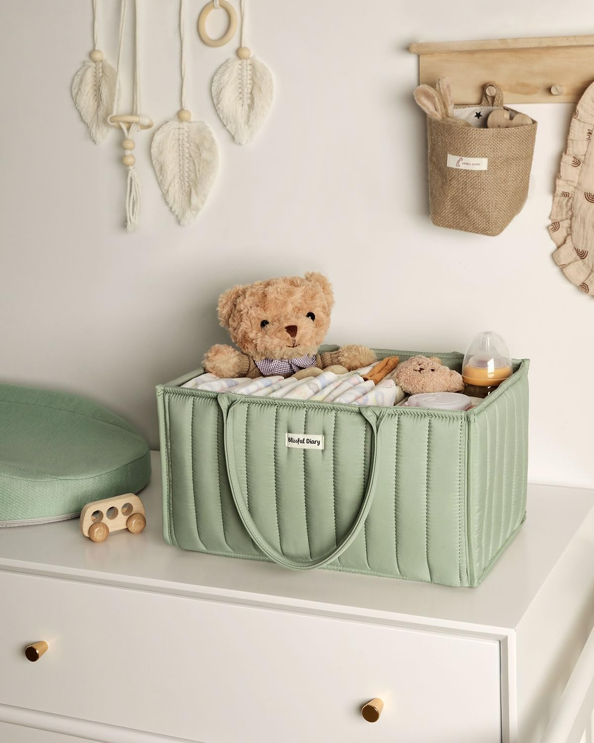 Nursery decor featuring storage basket on dresser with teddy bear, striped blanket, and baby essentials. Wall decor includes feathers and a pouch