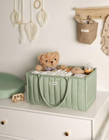 Nursery decor featuring storage basket on dresser with teddy bear, striped blanket, and baby essentials. Wall decor includes feathers and a pouch