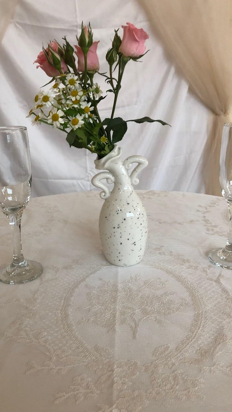 Porcelain vase with floral plan  holds pinkish  roses and daisies connected  an elegant lace-draped table, flanked by 2  bare  vino  glasses
