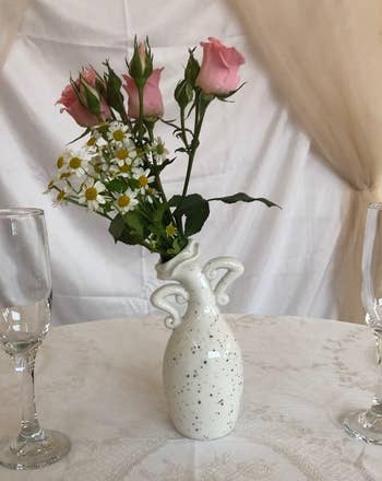 Porcelain vase with floral plan holds pinkish roses and daisies connected an elegant lace-draped table, flanked by 2 bare vino glasses