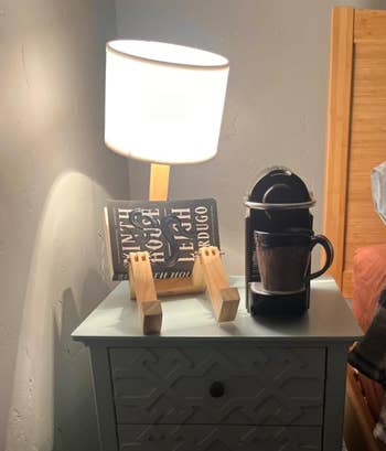 A bedside array  with a lamp, a publication  connected  a woody  stand, and a single-serve java  shaper  with a mug