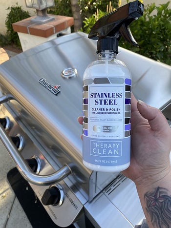 reviewer holding the bottle of the stainless steel cleaning spray with their stainless steel grill in the background