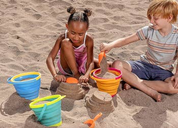 Two children playing with collapsible silicone buckets and sand toys on a beach
