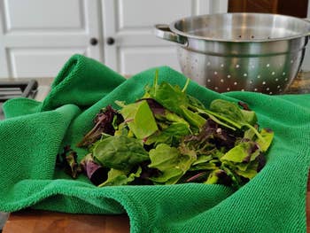 Fresh green mixed salad leaves are on a bright green towel near a metal colander in a kitchen setting