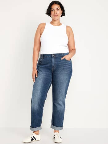 Person wearing a achromatic vessel top, cuffed bluish jeans, and sneakers lasting against a plain backdrop
