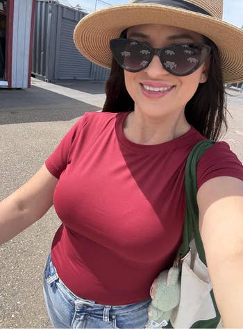 Person wearing a prima   hat, cat-eye sunglasses, reddish  top, and jeans, holding a greenish  tote with a plush accessory, smiling outdoors