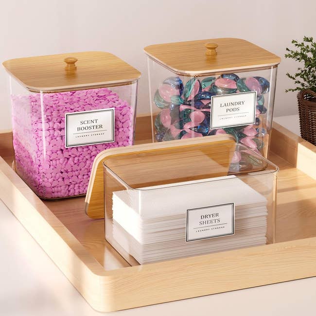 Three labeled containers hold pink scent boosters, colorful laundry pods, and white dryer sheets, displayed on a wooden tray