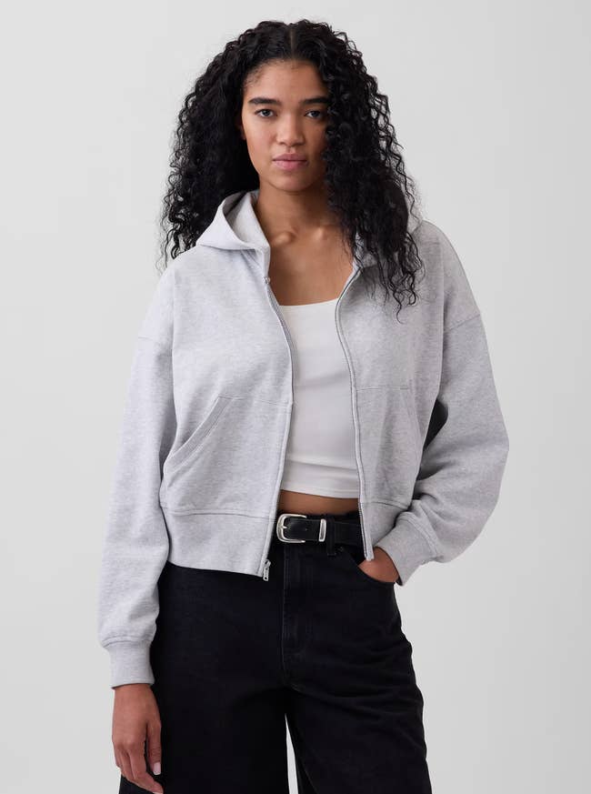 Person wearing a hooded zip-up overgarment   implicit    a achromatic  garment  and achromatic  pants, lasting  against a plain backdrop