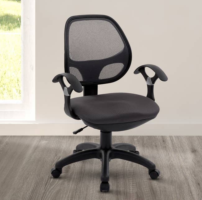 An ergonomic office chair with a mesh back, cushioned seat, and adjustable armrests on a wooden floor in a bright room