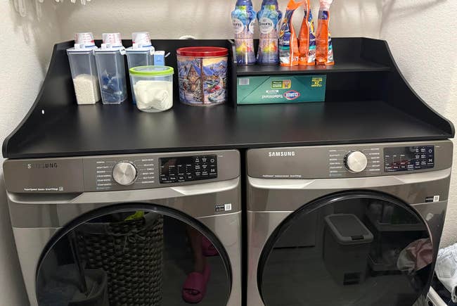 Laundry country with Samsung washer and dryer. Shelf holds detergent, cleaning supplies, and retention containers. Nearby laundry handbasket is visible