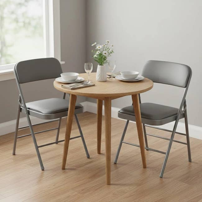 A round wooden dining table with two place settings, accompanied by two gray folding chairs, set in a well-lit room by a window