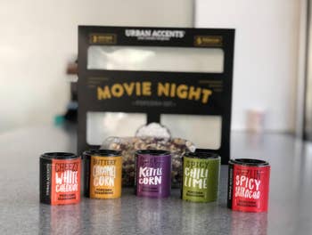 Reviewer image of product box open with three bags of kernels behind the orange, yellow, purple, green, and red small seasoning containers