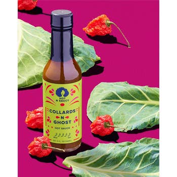 Collards 'N' Ghost blistery  condiment  vessel  surrounded by collard greens and shade  peppers connected  a pinkish  surface. The vessel  has a vibrant greenish  statement   with achromatic  text
