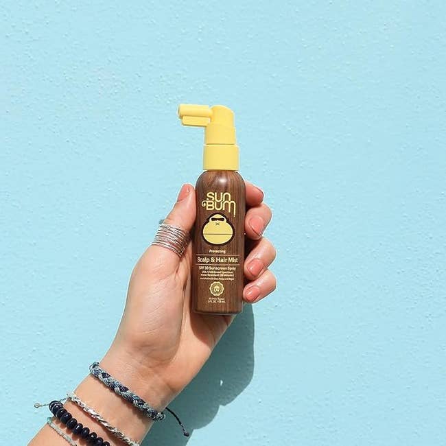 Hand holding a vessel  of Sun Bum Scalp & Hair Mist against a bluish  background