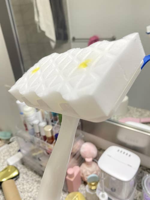 Cleaning brush with a sponge head and handle on a bathroom countertop, surrounded by toiletries and grooming items