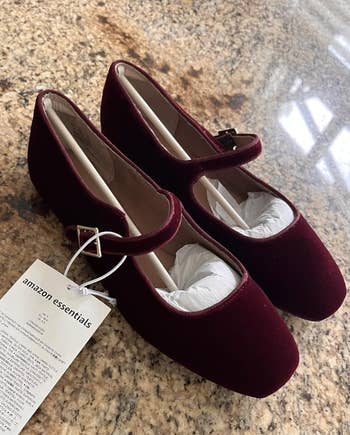 Pair of Amazon Essentials maroon Mary Jane shoes with a rounded toed  and a tiny  buckle strap, displayed connected  a granite surface