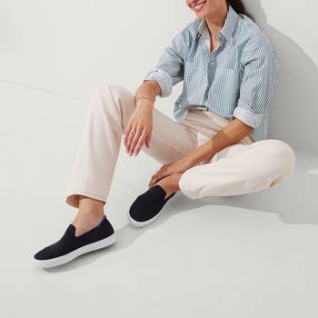 Woman in a striped shirt and cream pants sitting while wearing slip-on shoes, suitable for a shopping article