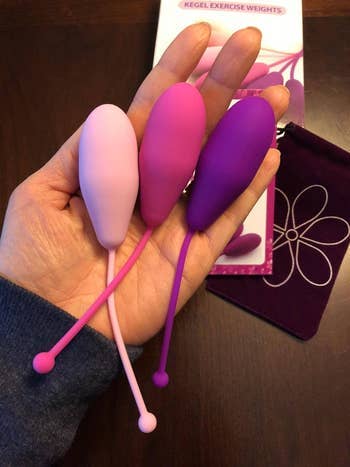 Hand holding three kegel exercise weights of varying shapes and sizes on a table with a floral pouch