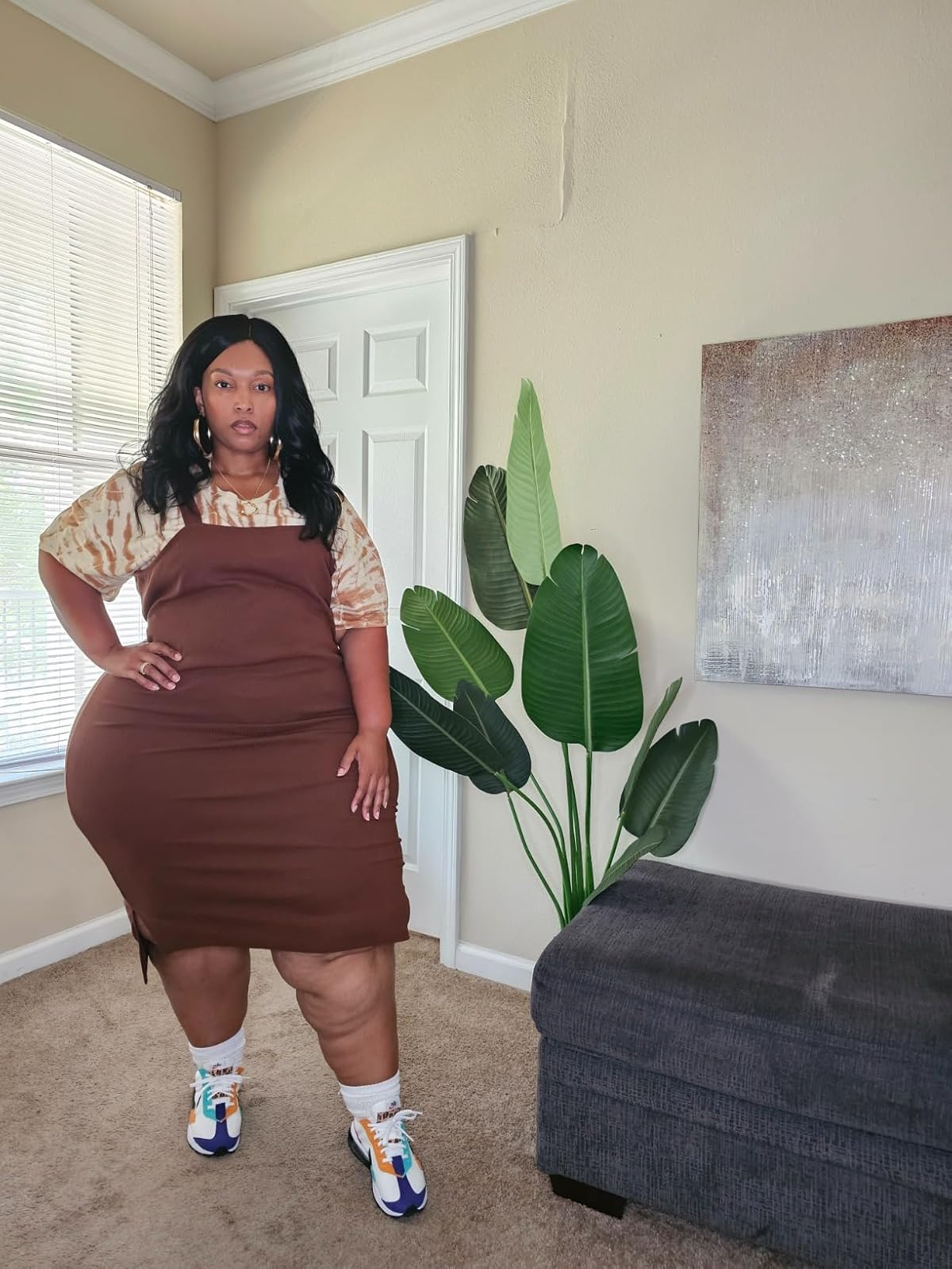 Person poses indoors wearing a brown dress layered over a tie-dye shirt, with colorful sneakers. They stand next to a potted plant and a textured wall art