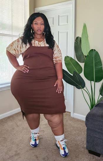Person poses indoors wearing a brown dress layered over a tie-dye shirt, with colorful sneakers. They stand next to a potted plant and a textured wall art