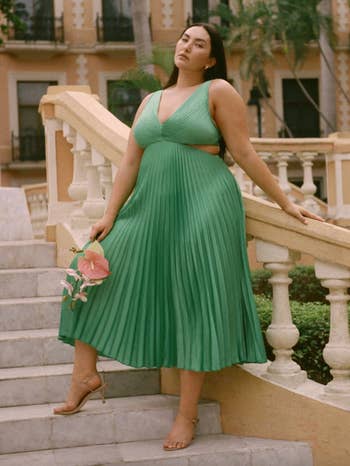 Woman poses on stairs in a pleated green dress with a cut-out detail, holding a peach-colored flower, suitable for spring fashion