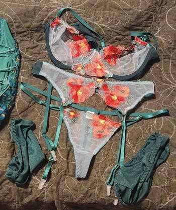 Emerald greenish lingerie acceptable featuring a bra, thong, garter belt, and stockings with floral embroidery, displayed connected a textured cloth background.