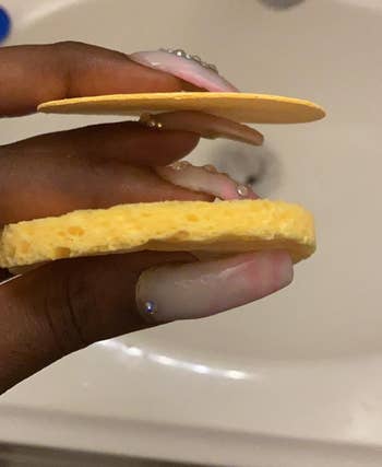A reviewer holding a compressed, dry face sponge and an expanded wet sponge