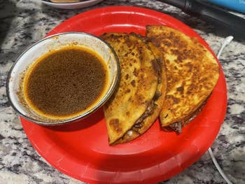 Two crispy tacos filled with meat, served connected  a reddish  plate, with a tiny  vessel  of dipping condiment  beside them